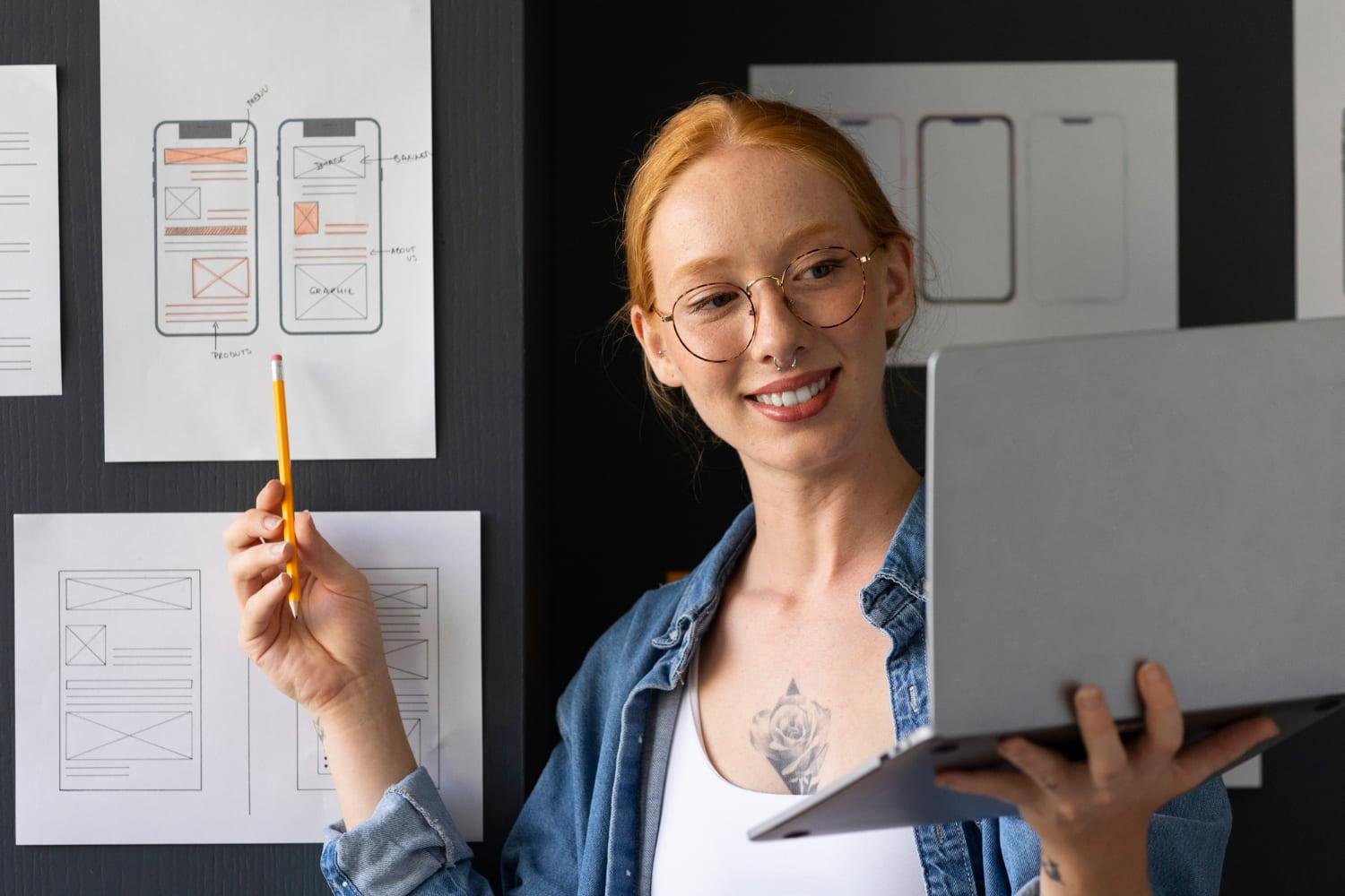 Mulher branca com laptop na mão esquerda, apontando para quadro com lápis na mão direita